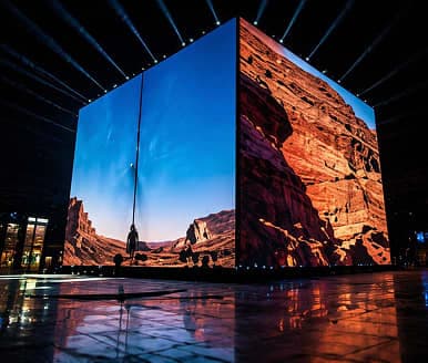 Large luminous digital cube installation at night with desert and rock imagery on its screens, reflective floor and upward light beams